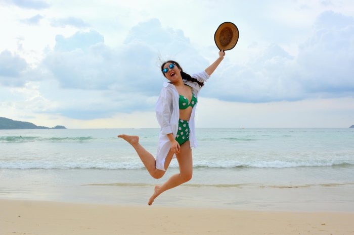 mujer con pelo recogido saltando en la playa con sombrero en la mano